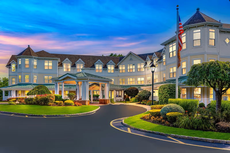 Exterior view of a large, elegant senior living facility building at dusk with well-maintained landscaping, an American flag on a flagpole, and a covered entrance driveway.