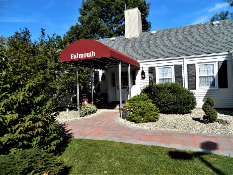 Exterior view of a single-story building with a gray shingled roof and white siding. There is a red canopy over the entrance with the word 'Falmouth' written on it. The entrance is surrounded by bushes, small trees, and a paved walkway leading up to the door. The sky is clear and blue.