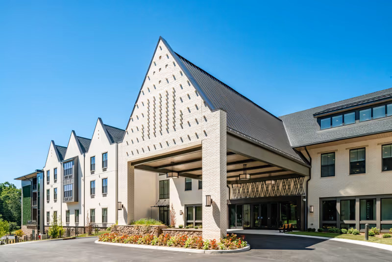 Exterior view of a modern senior living facility building with a large covered entrance, white brick walls, multiple windows, and a clear blue sky.