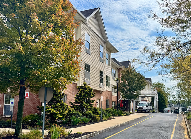 Three-story assisted living building front with landscaping, a tree-lined sidewalk, and a driveway with a parked van.