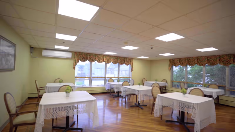 A dining room with several tables covered in white lace tablecloths, each table having a small potted plant centerpiece. The room has wooden floors, large windows with patterned valances, and multiple chairs around the tables. The ceiling has recessed lighting panels.