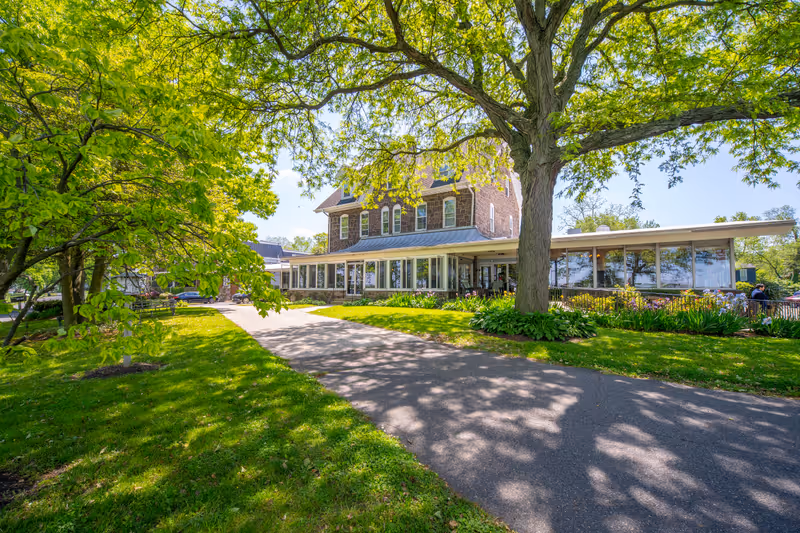 A large, multi-story brick building with a metal roof surrounded by lush green trees and a well-maintained lawn. A paved pathway leads up to the building, which has large windows and a porch area. The scene is bright and sunny with shadows from the trees cast on the ground.