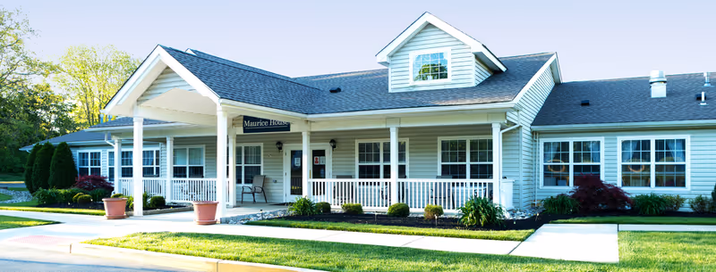 Exterior front view of Maurice House Assisted Living Community, a single-story building with light-colored siding, a covered porch with white railings, several windows, and a small dormer on the roof. The surrounding area includes green grass, shrubs, and trees.