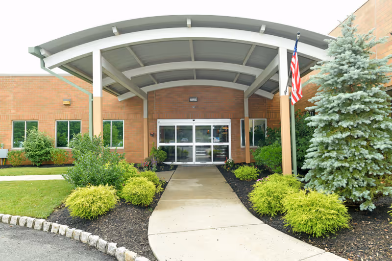 Entrance to a brick building with a covered walkway supported by white beams. There are landscaped bushes and a small tree on either side of the concrete path leading to automatic glass doors. An American flag is mounted on the right side near the entrance.