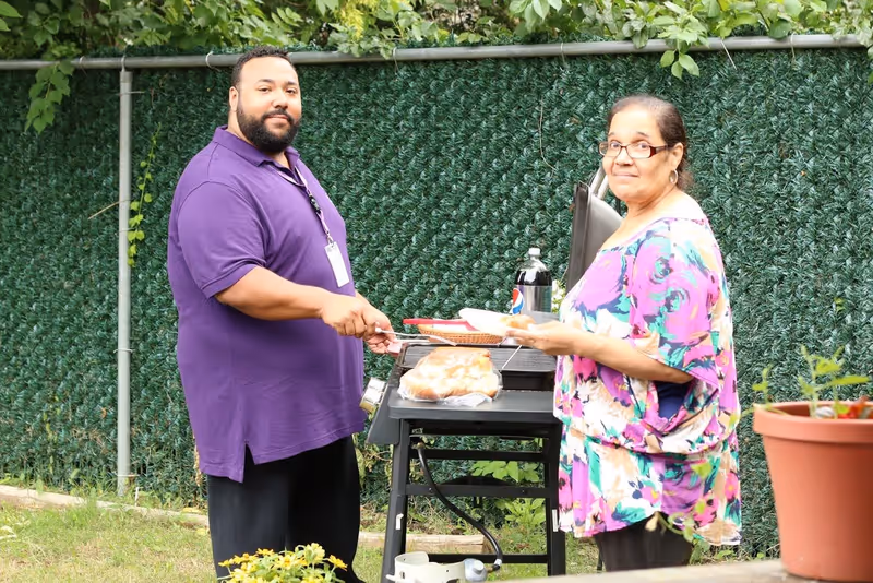 A man and a woman standing outdoors next to a grill. The man is wearing a purple shirt and holding a spatula, while the woman is wearing glasses and a colorful floral top. There is a bottle of soda and a pack of buns on the grill. They are in a garden area with green foliage and a fence in the background.