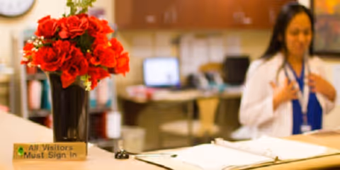 Reception desk with a vase of red flowers and a sign that reads 'All Visitors Must Sign In.' In the background, a woman in a white coat is standing in an office or medical facility setting.