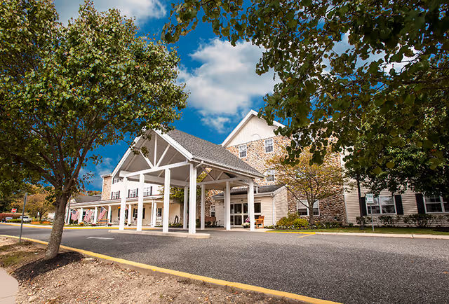Front exterior of a three-story senior living building with a covered entrance/porte-cochere, trees, and driveway.