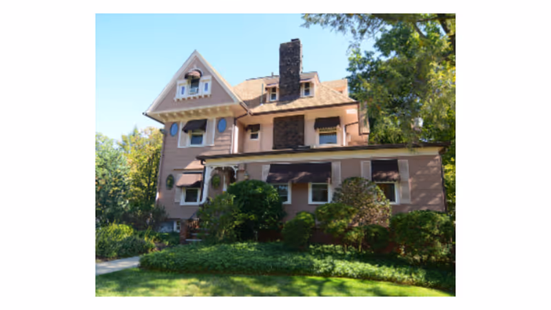 Exterior view of a large, multi-story residential building with a steep roof, multiple windows with awnings, a central chimney, and surrounded by green trees and bushes under a clear blue sky.