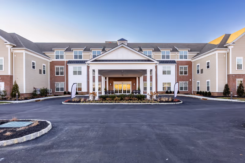 Front exterior view of a large, three-story senior living facility building with beige siding and red brick accents. The building has multiple windows and a covered entrance with white pillars. There is a circular driveway and landscaped area in front of the entrance under a clear blue sky.