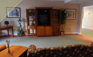 Interior view of a living room in Heritage Assisted Living featuring a floral patterned sofa, a wooden coffee table with a small flower vase, a large wooden entertainment center with a TV, potted plants, framed artwork on the walls, and a hallway leading to other rooms.