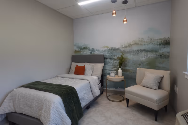 A small, cozy bedroom with a single bed made with white bedding and a green throw blanket. There is a gray upholstered headboard and a red accent pillow. Next to the bed is a round wooden side table with a white vase holding green plants and a small picture frame. A beige armchair with a matching pillow sits beside the table. The wall behind the bed features a calming mural with abstract green and blue tones. Two pendant lights hang from the ceiling.