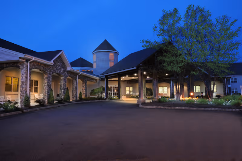 Exterior view of Crane's Mill facility at dusk, showing a building with stone columns, a covered entrance, and a landscaped area with trees and flowers under a clear blue sky.