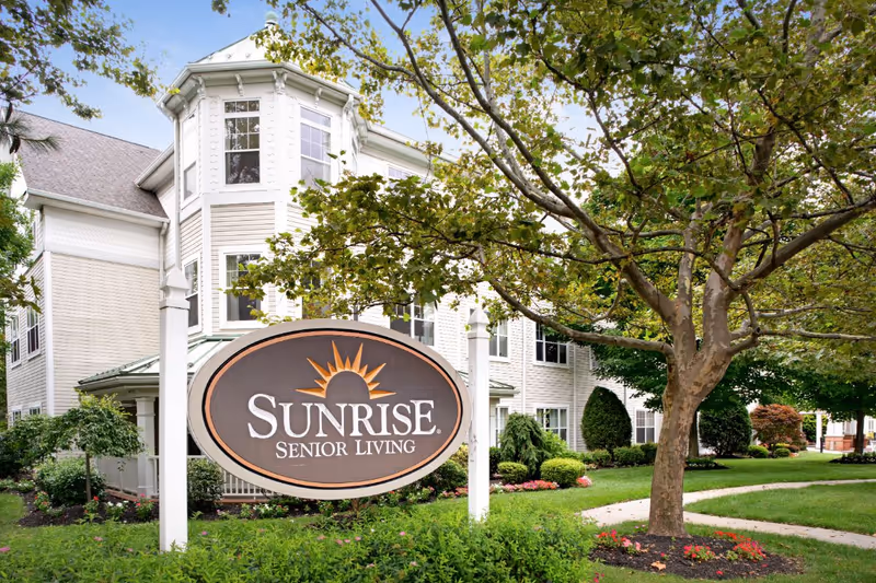 Large Sunrise Senior Living sign in front of the facility's landscaped building exterior and trees.