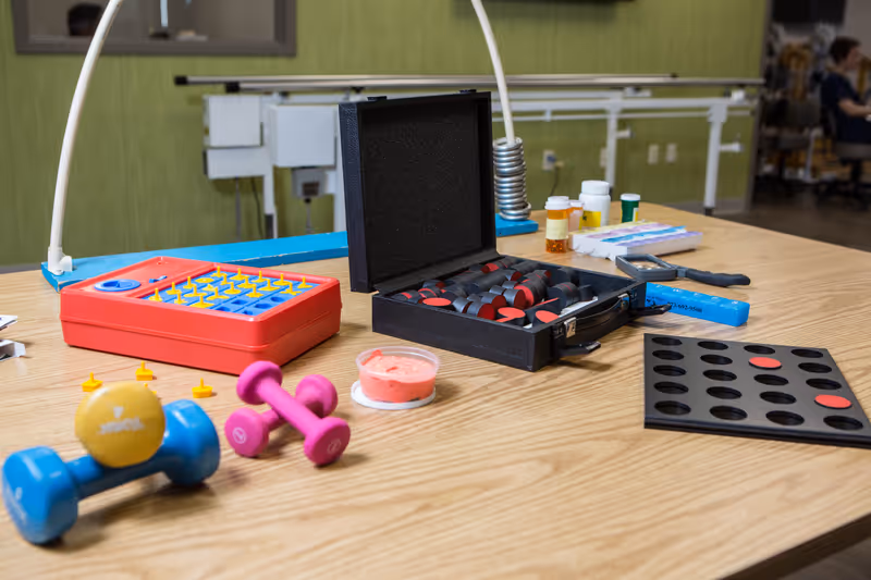 A wooden table with various therapy and exercise tools including colorful plastic pegs, a black case with red and black checkers pieces, small dumbbells, pill bottles, and a Connect Four game board. The background shows a green wall and part of a therapy or activity room.
