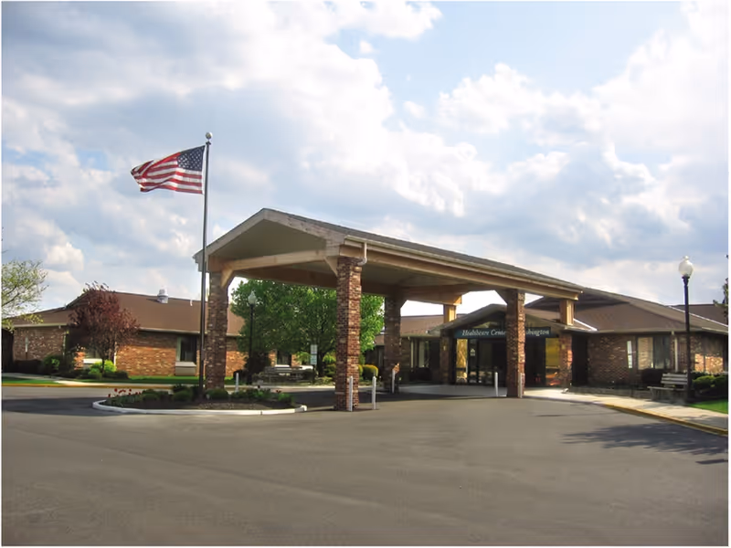 Exterior view of the Center for Rehabilitation & Nursing at Washington Township showing a brick building with a covered entrance, an American flag on a flagpole, and a partly cloudy sky.