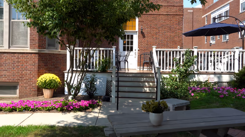 Outdoor patio area of a senior living facility with a brick building in the background. There are stairs leading up to a white railing deck with chairs and a table. Flower beds with pink and yellow flowers surround the patio. A picnic table with a small potted plant is in the foreground, and a large umbrella provides shade on the right side.