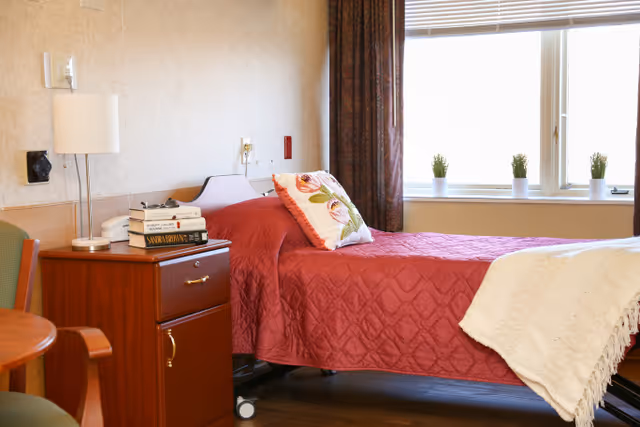 A single bed with a red quilt and a floral pillow in a well-lit room. Next to the bed is a wooden nightstand with a lamp and a stack of books. A window with blinds and small potted plants on the sill is visible in the background.