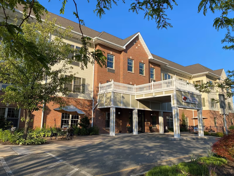 Front entrance of a multi-story brick-and-siding senior living building with a covered porte-cochère, balcony, and landscaped grounds under a clear blue sky.