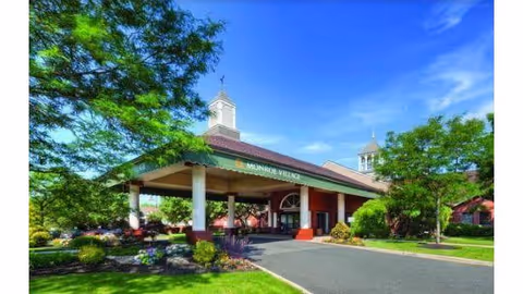 Entrance of Monroe Village senior living facility with a covered driveway, surrounded by green trees and landscaped bushes under a bright blue sky.