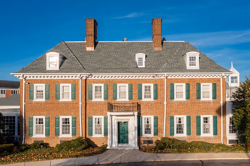 Front exterior view of a large brick building with green shutters and a gray shingled roof under a clear blue sky. The building has two chimneys and a central entrance with a small balcony above it. There are bushes and a sidewalk in front of the building.