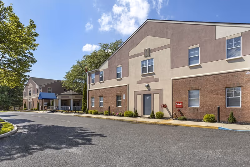 Exterior view of a two-story senior living facility building with a combination of brick and beige siding. The building has several windows and a main entrance with a blue awning. There are trees and shrubs along the sidewalk and a clear blue sky with some clouds.