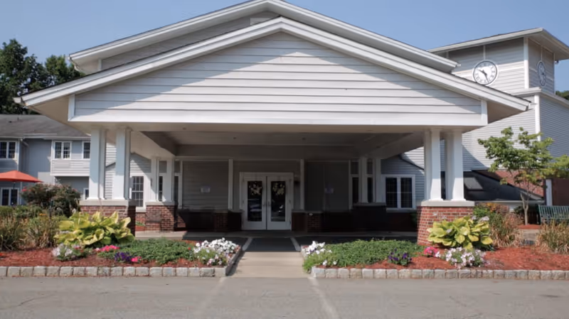 Front entrance of a senior living facility with a covered drop-off area supported by white columns. The building has light gray siding with brick accents at the base and a clock tower visible on the right side. There are landscaped flower beds with green plants and colorful flowers on either side of the entrance walkway.