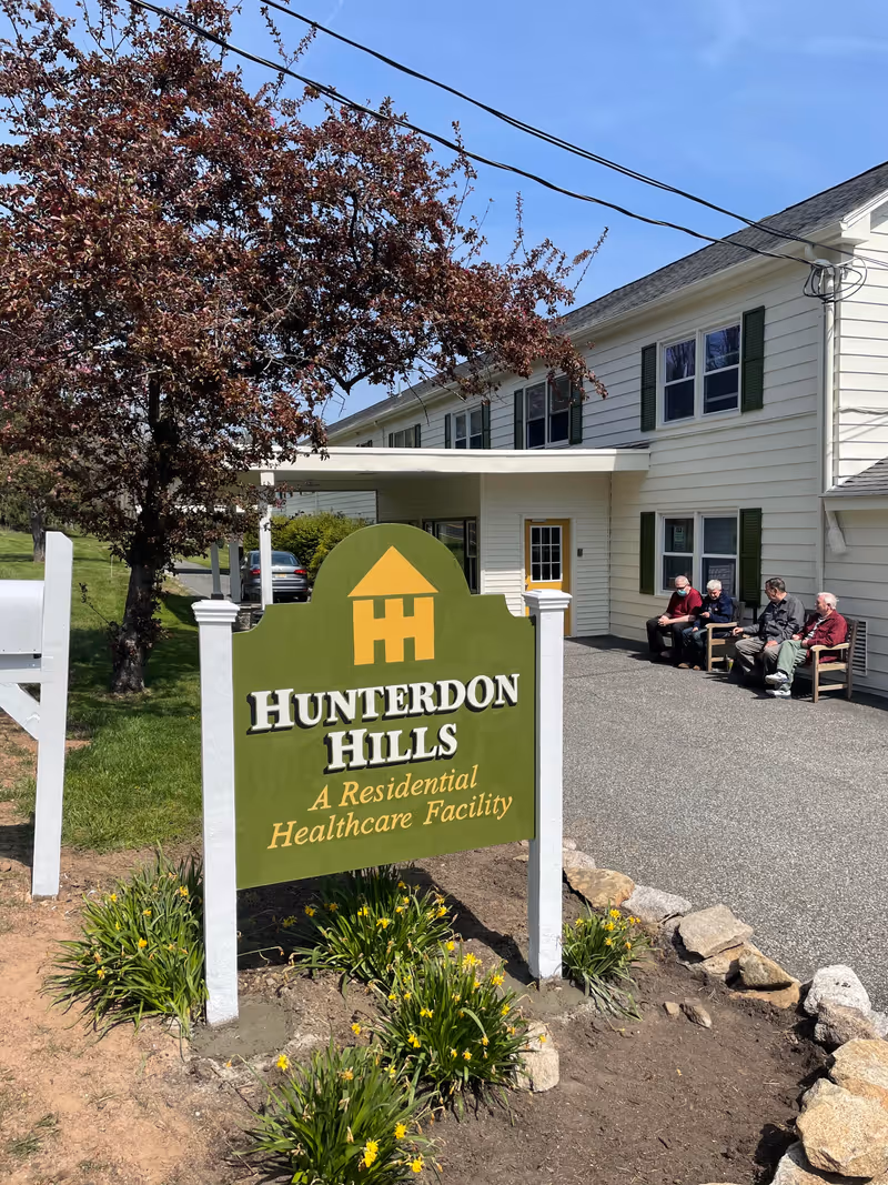 Outdoor view of Hunterdon Hills Residential Healthcare Facility sign with a green and yellow logo. The building is white with green shutters, and four elderly people are sitting on benches near the entrance. There is a tree with reddish leaves and some yellow flowers planted around the sign.