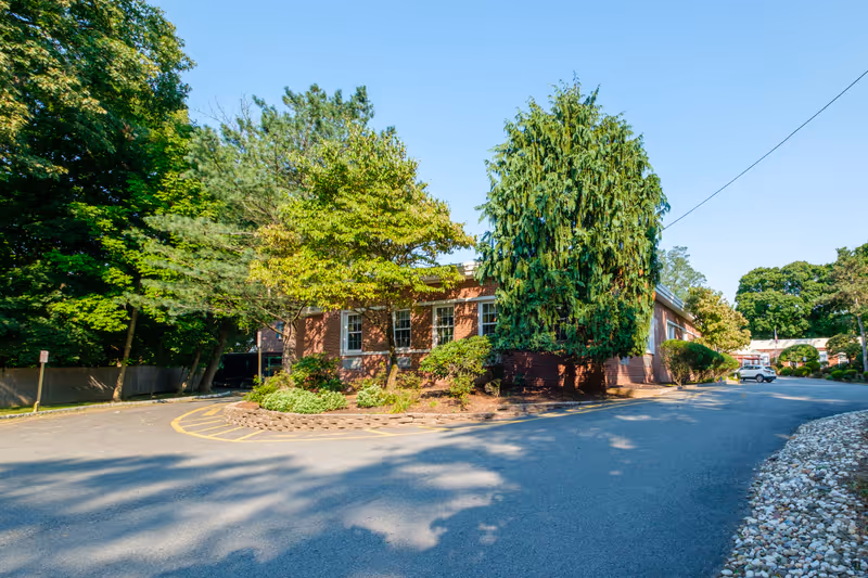 Brick single-story building surrounded by trees and landscaping with a curved driveway and a parked car.