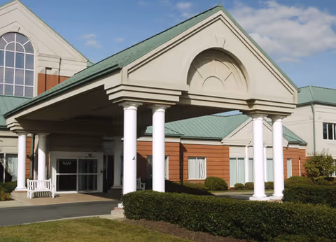 Exterior view of Atrium Senior Living Of Princeton showing the main entrance with a covered driveway supported by white columns, a green roof, and brick and beige walls. There are bushes and a lawn in front of the building under a partly cloudy sky.