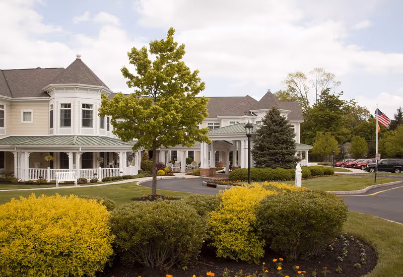 Exterior view of a senior living facility with a large, light-colored building featuring a covered porch and multiple windows. The foreground shows well-maintained landscaping with green and yellow bushes, a tree, and a curved driveway. An American flag is visible on the right side near parked cars.