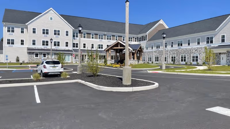 Exterior view of Ridgewood Senior Living at Historic Smithville showing a large, multi-story building with white siding and stone accents. The building has many windows and a covered entrance. In front is a paved parking lot with a few cars and landscaped areas with small trees and bushes.