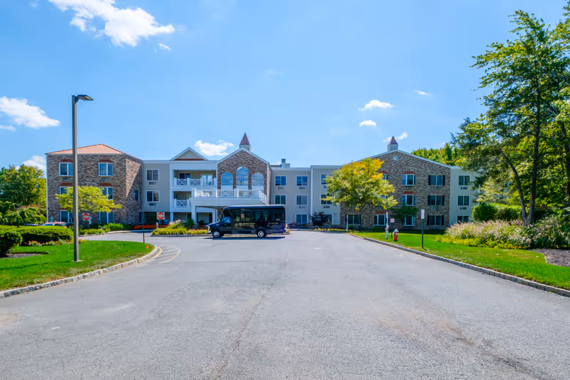Front exterior view of Brandywine Princeton by Monarch, a multi-story senior living facility with a mix of stone and light-colored siding, surrounded by green trees and landscaping under a blue sky with some clouds. A black shuttle van is parked in front of the building.