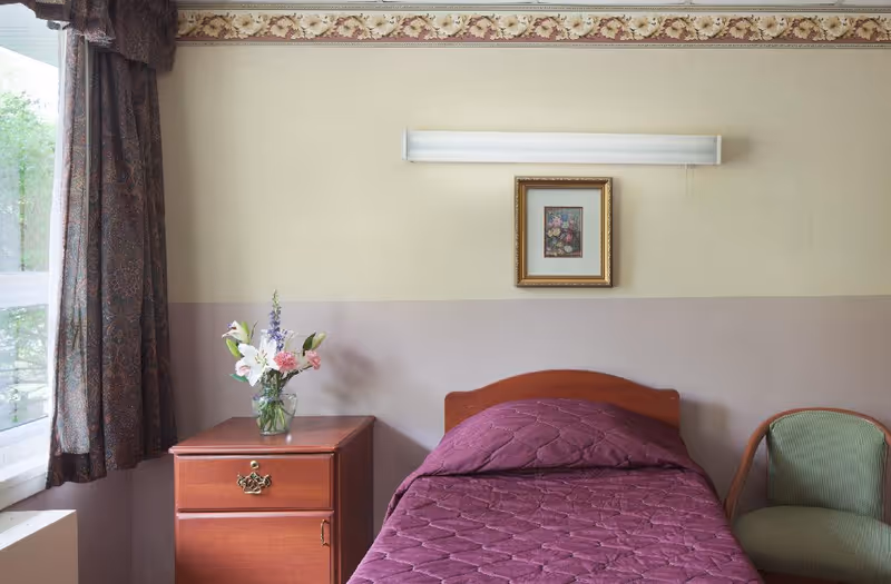 A single bed with a purple quilted bedspread next to a wooden nightstand with a glass vase of flowers. There is a green upholstered chair to the right of the bed. A framed floral picture hangs on the wall above the bed, and a window with patterned curtains is on the left side of the room.