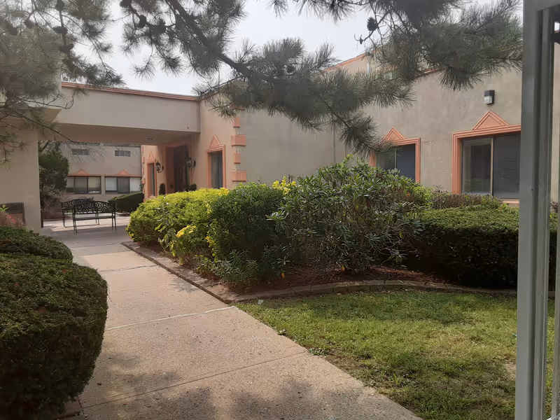 Outdoor courtyard area of a building with a paved walkway, green bushes, and trees. The building has beige walls with orange trim around the windows and doors. There is a metal bench visible under a covered walkway.