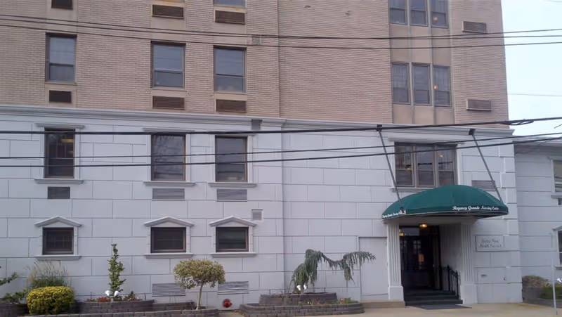 Exterior view of a multi-story building with beige and white walls, several windows, and a green awning above the entrance that reads 'Regency Grande Nursing Center'. There are small landscaped areas with bushes and small trees in front of the building.