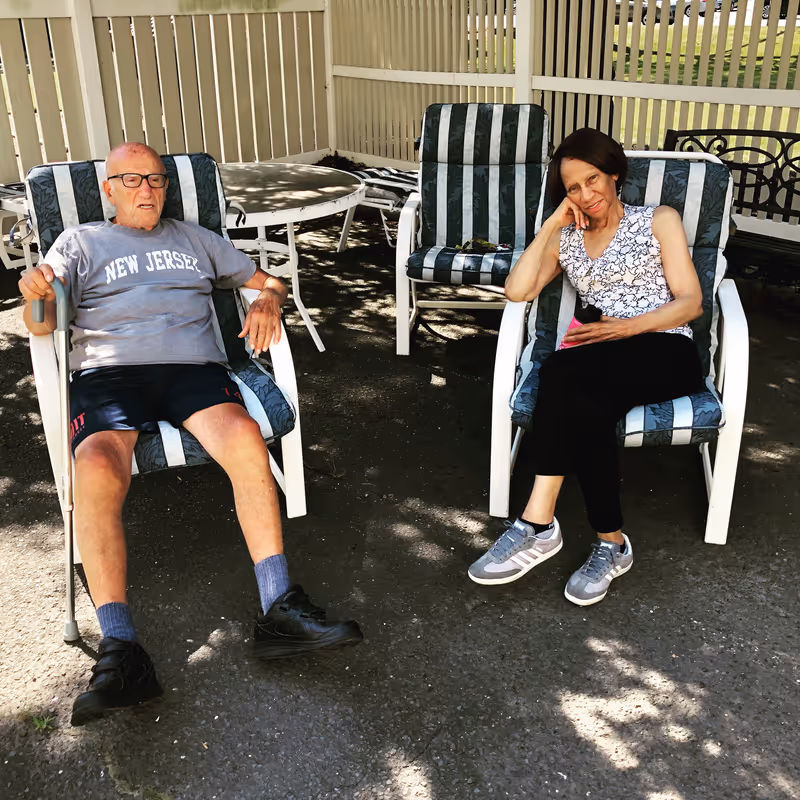 An elderly man and woman sitting outdoors on striped cushioned chairs under a shaded patio area. The man is wearing a gray New Jersey t-shirt, black shorts, and black shoes, holding a cane. The woman is wearing a sleeveless patterned top, black pants, and gray sneakers, resting her head on her hand. Behind them is a white round table and additional chairs with a white fence in the background.