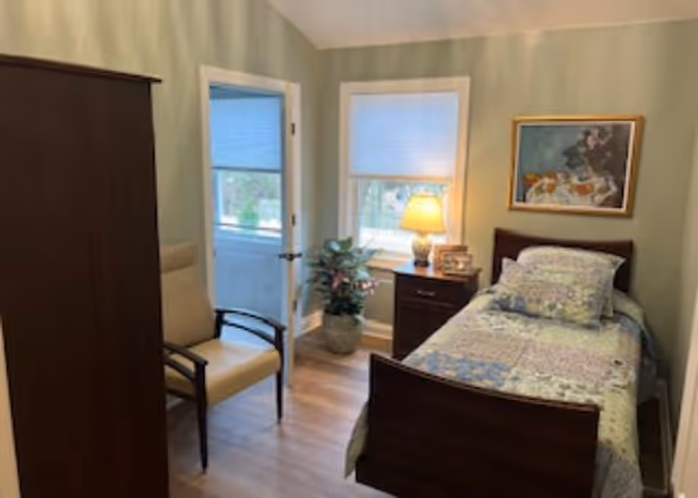 Small furnished bedroom with a single bed, nightstand and lamp, chair, wardrobe and windows.