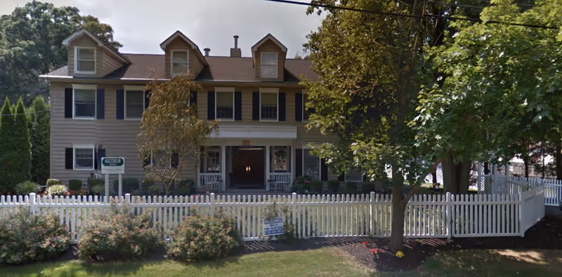 A large two-story beige house with black shutters and three dormer windows on the roof, surrounded by a white picket fence and trees. There is a sign in front of the house and a 'Hiring' sign on the fence.