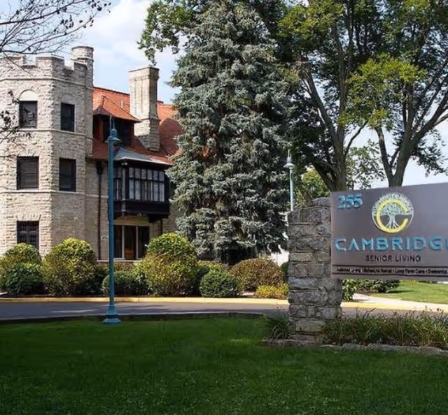 Exterior view of Cambridge Enhanced Senior Living facility showing a stone building with a turret, surrounded by trees and bushes, with a sign in the foreground displaying the facility name and address number 255.