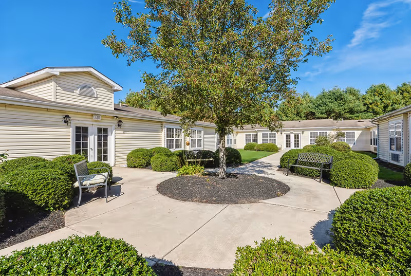 Outdoor courtyard area of a senior living facility with a central tree surrounded by bushes and benches. The courtyard is paved with concrete walkways and is bordered by single-story beige buildings with white-framed windows and doors under a clear blue sky.