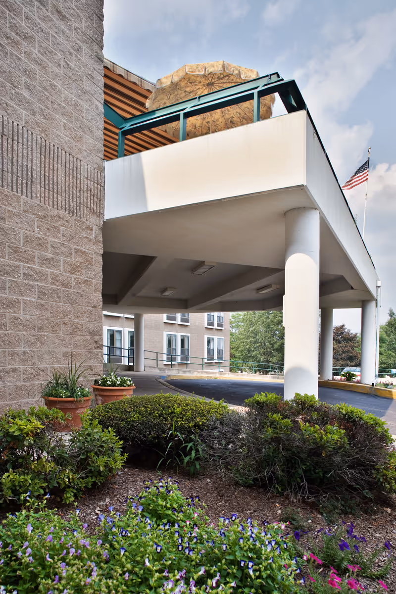 Exterior view of a building entrance with a covered driveway supported by white columns. There are green shrubs and flowering plants in the foreground, and an American flag is visible on a flagpole to the right. The sky is partly cloudy.