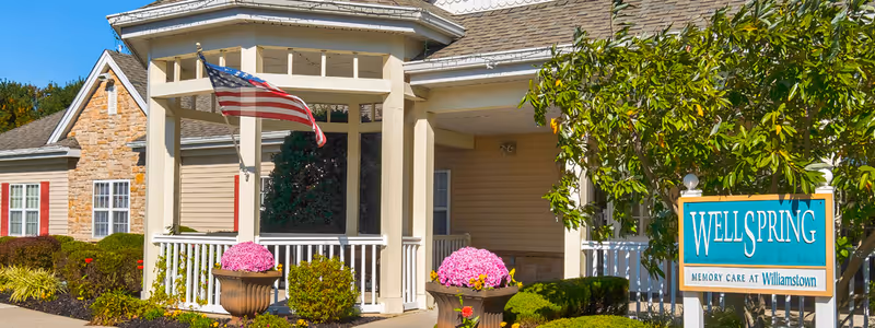 Exterior view of a senior living facility entrance with a covered porch, American flag, stone and beige siding walls, and landscaping including bushes and flowers. A sign reads 'Wellspring Memory Care at Williamstown'.