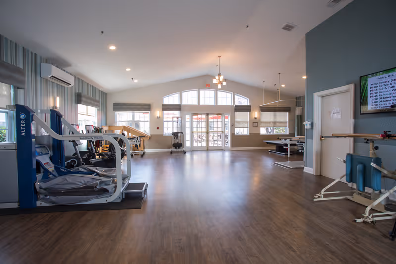 A spacious rehabilitation or physical therapy room with exercise equipment including an anti-gravity treadmill, parallel bars, and other therapy apparatus. The room has large windows and glass doors letting in natural light, wood flooring, and soft wall colors with striped wallpaper on one side.