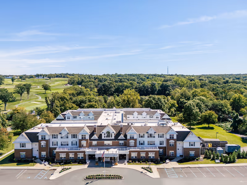 Aerial view of the front of a large three-story senior living building with a parking lot, surrounding trees, and a golf course in the background.