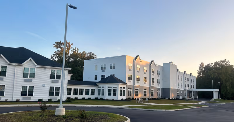 Exterior view of a large, modern senior living facility building with white siding and multiple windows, surrounded by a paved driveway and landscaped grass areas under a clear sky at sunset.