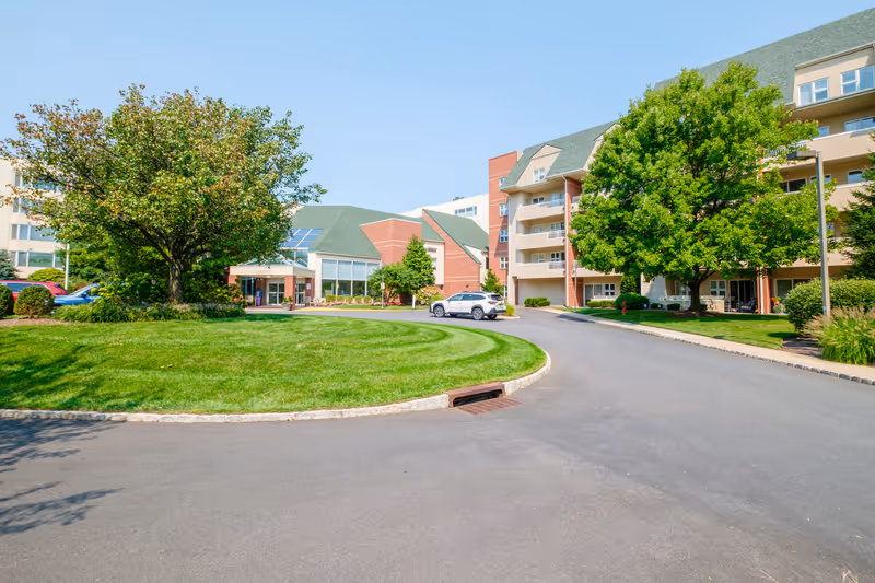 Front driveway and landscaped lawn leading to the entrance of a multi-story senior living building with trees and parked cars.
