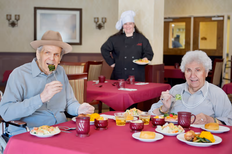 Two elderly residents seated at a table with plates of food in a senior care dining room while a chef stands smiling behind them.