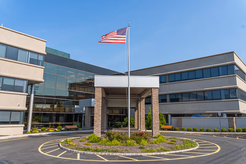 Front entrance of a modern senior living facility with a flagpole, circular driveway, and landscaped island.