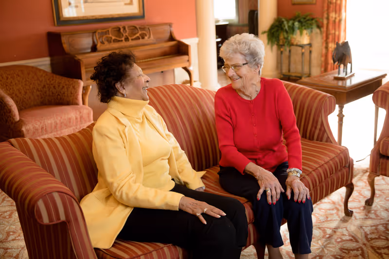 Two elderly women sitting on a striped sofa in a warmly decorated living room, smiling and engaging in conversation. Behind them is a wooden piano and a framed picture on the wall. The room has a cozy and inviting atmosphere with warm colors and comfortable furniture.