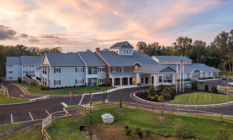 Exterior view of HarborChase Of Princeton senior living facility at sunset, showing a large multi-story building with a covered entrance, surrounding parking lot, landscaped greenery, and American flags near the entrance.